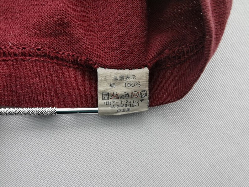 May include: Close-up of a burgundy fabric with a sewn seam. A rectangular tag is attached, displaying Japanese text and symbols, including "100%" and care instructions. The tag is white with black text.
