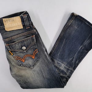 Op de afbeelding: Verbleekte blauwe denim jeans met oranje stiksels op de achterzakken. De jeans hebben een distressed look en een leren patch met de merknaam "EDWIN". De jeans zijn opgevouwen en op een wit oppervlak weergegeven.