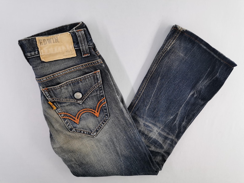 Op de afbeelding: Verbleekte blauwe denim jeans met oranje stiksels op de achterzakken. De jeans hebben een distressed look en een leren patch met de merknaam "EDWIN". De jeans zijn opgevouwen en op een wit oppervlak weergegeven.
