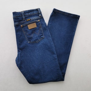 May include: Dark blue denim jeans with a classic five-pocket design. Features a brown leather patch and the Wrangler logo on the back pocket. The jeans are folded neatly, showcasing the straight leg cut.