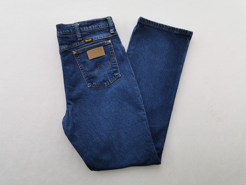 May include: Dark blue denim jeans with a classic five-pocket design. Features a brown leather patch and the Wrangler logo on the back pocket. The jeans are folded neatly, showcasing the straight leg cut.