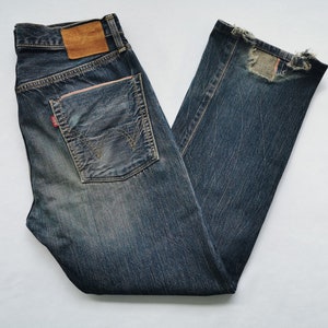 Pu&ograve; includere: Jeans in denim blu scuro con un aspetto invecchiato. Presentano una toppa in pelle marrone sulla cintura posteriore e un'etichetta rossa sulla tasca posteriore. I jeans hanno un aspetto sbiadito e una toppa sulla gamba destra.