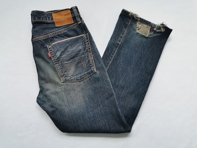 Pu&ograve; includere: Jeans in denim blu scuro con un aspetto invecchiato. Presentano una toppa in pelle marrone sulla cintura posteriore e un'etichetta rossa sulla tasca posteriore. I jeans hanno un aspetto sbiadito e una toppa sulla gamba destra.