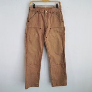 May include: Tan work pants with reinforced knees and multiple pockets hang on a wooden hanger. The pants are made of a durable fabric and feature double stitching. The pants are a neutral color, suitable for various outfits.
