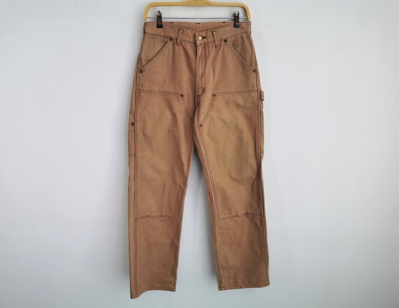 May include: Tan work pants with reinforced knees and multiple pockets hang on a wooden hanger. The pants are made of a durable fabric and feature double stitching. The pants are a neutral color, suitable for various outfits.