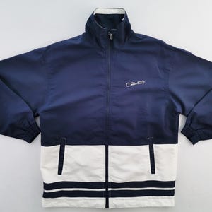 Puede incluir: Chaqueta con cremallera azul marino y blanca. La chaqueta tiene cuello alto, dos bolsillos delanteros y el texto "C. Rich Sport" bordado en el pecho. La mitad inferior de la chaqueta es blanca con dos rayas azul marino.