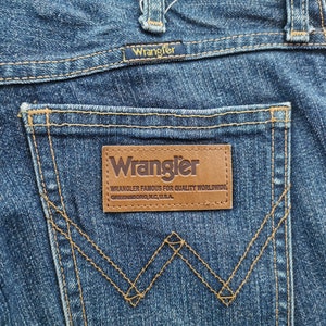 May include: Close-up of blue denim jeans featuring the Wrangler logo on the back pocket and waistband. The jeans have classic stitching details and a brown leather patch with the brand's name and location.