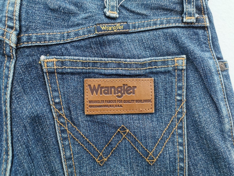 May include: Close-up of blue denim jeans featuring the Wrangler logo on the back pocket and waistband. The jeans have classic stitching details and a brown leather patch with the brand's name and location.