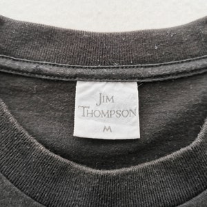 May include: Close-up of a black t-shirt collar with a white tag. The tag reads "Jim Thompson" in a serif font, with the size "M" printed below. The shirt's fabric appears to be a soft cotton material.
