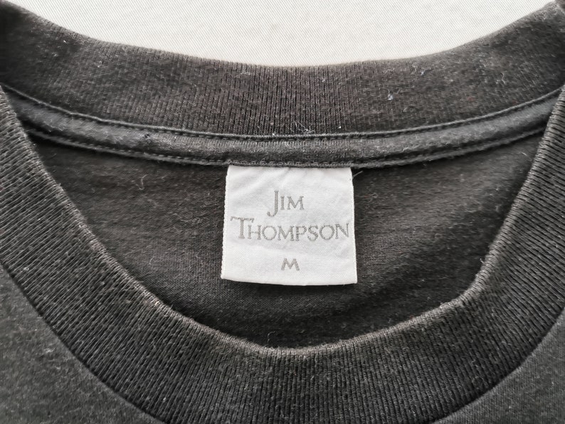May include: Close-up of a black t-shirt collar with a white tag. The tag reads "Jim Thompson" in a serif font, with the size "M" printed below. The shirt's fabric appears to be a soft cotton material.