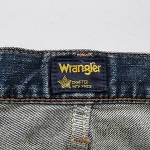 May include: Close-up of a pair of blue denim jeans. The Wrangler label is visible, featuring the brand name in gold lettering on a navy blue background, with a star graphic and the text "Crafted with Pride".