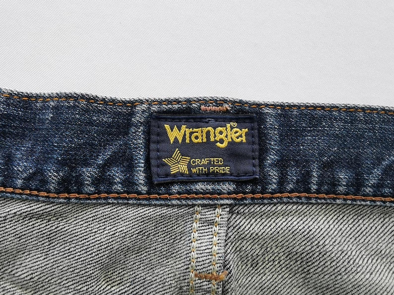 May include: Close-up of a pair of blue denim jeans. The Wrangler label is visible, featuring the brand name in gold lettering on a navy blue background, with a star graphic and the text "Crafted with Pride".