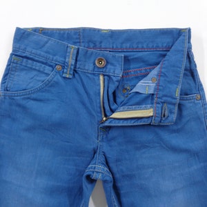 K&ouml;nnte beinhalten: Nahaufnahme einer blauen Jeans mit Knopfverschluss und Rei&szlig;verschluss. Die Jeans hat gelbe N&auml;hte und rote N&auml;hte auf der Innenseite. Die Taschen sind sichtbar, und die Jeans liegt flach auf einer wei&szlig;en Oberfl&auml;che.