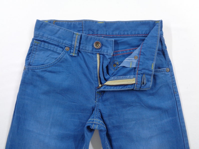 K&ouml;nnte beinhalten: Nahaufnahme einer blauen Jeans mit Knopfverschluss und Rei&szlig;verschluss. Die Jeans hat gelbe N&auml;hte und rote N&auml;hte auf der Innenseite. Die Taschen sind sichtbar, und die Jeans liegt flach auf einer wei&szlig;en Oberfl&auml;che.