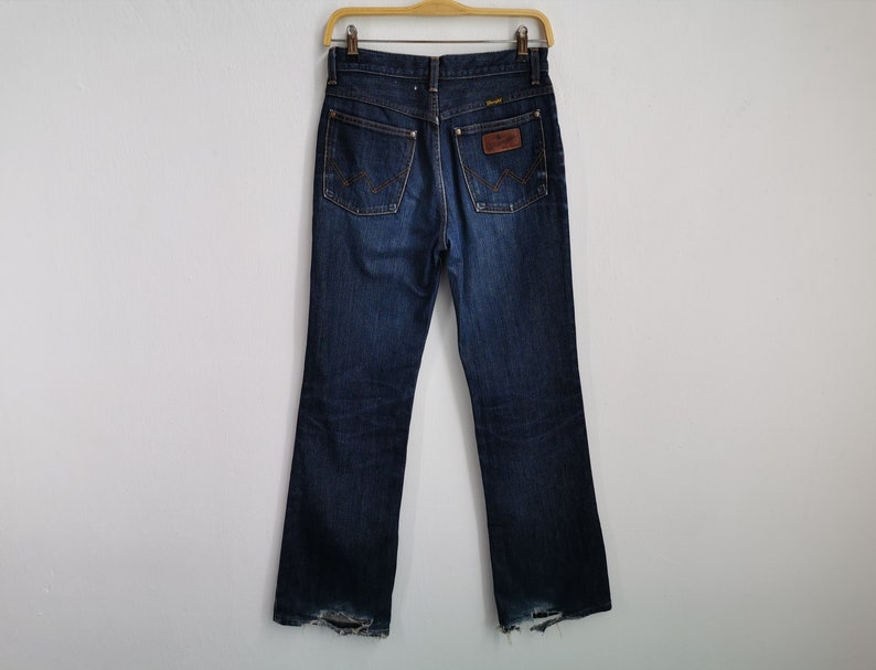 May include: Dark wash denim jeans with a classic five-pocket design, hanging on a wooden hanger. The jeans feature a brown leather patch with the brand name and a faded, worn look at the bottom of the legs.
