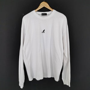 Op de afbeelding: Wit shirt met lange mouwen, een ronde hals en een klein zwart kangoeroelogo op de borst. De merknaam staat in kleine tekst rond de kraag. Het shirt hangt aan een houten kleerhanger.
