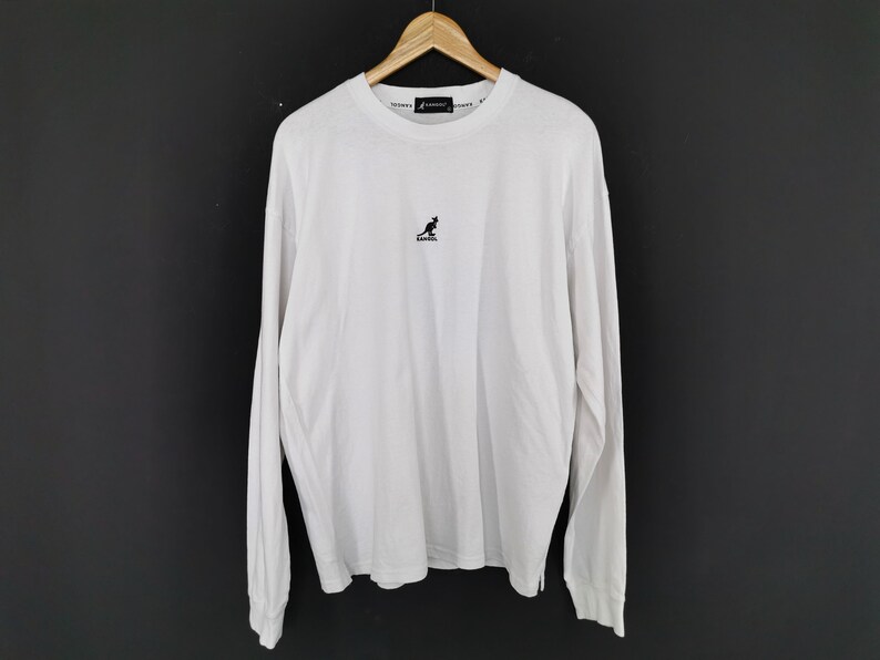 Op de afbeelding: Wit shirt met lange mouwen, een ronde hals en een klein zwart kangoeroelogo op de borst. De merknaam staat in kleine tekst rond de kraag. Het shirt hangt aan een houten kleerhanger.