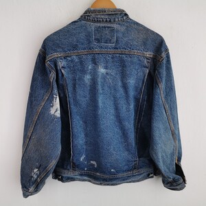May include: A vintage denim jacket in a medium wash blue, with a classic collar and button-up front. The jacket shows signs of wear, with faded patches and distressed areas, giving it a worn-in look. The back features a small, square label.