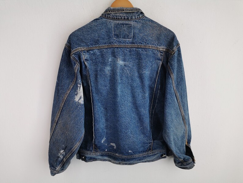 May include: A vintage denim jacket in a medium wash blue, with a classic collar and button-up front. The jacket shows signs of wear, with faded patches and distressed areas, giving it a worn-in look. The back features a small, square label.
