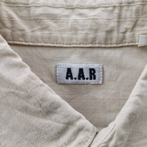 May include: A light beige shirt featuring a textured fabric and a collar. A rectangular white fabric label is sewn onto the inside neck area, displaying the black embroidered text A.A.R in a bold, sans-serif font with visible stitching.