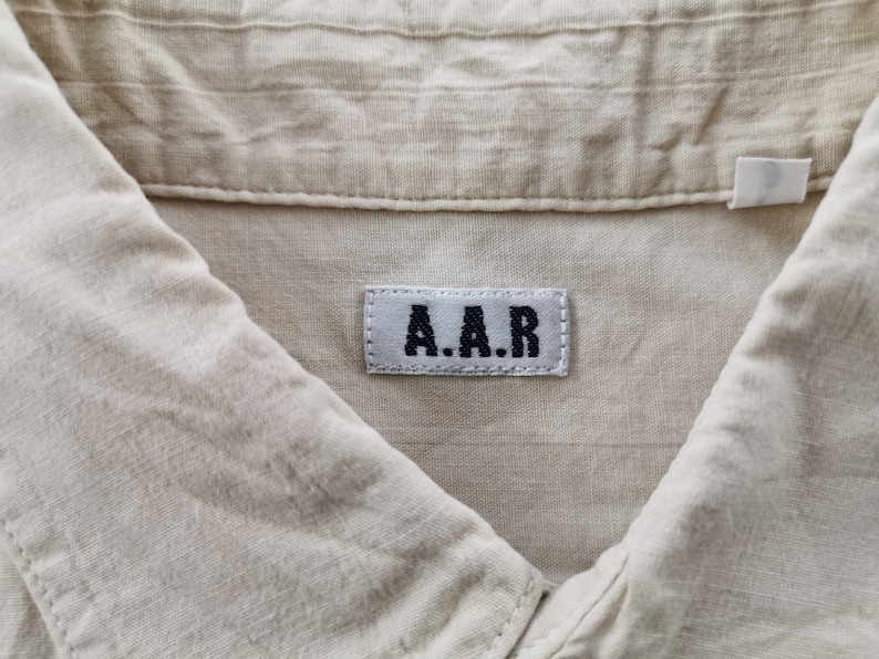 May include: A light beige shirt featuring a textured fabric and a collar. A rectangular white fabric label is sewn onto the inside neck area, displaying the black embroidered text A.A.R in a bold, sans-serif font with visible stitching.
