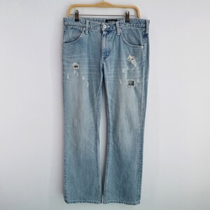 Pu&ograve; includere: Jeans in denim blu chiaro con dettagli strappati, tra cui sezioni strappate sul davanti. I jeans sono a gamba dritta e sono appesi a una gruccia di legno su sfondo bianco. Un classico capo d'abbigliamento casual.