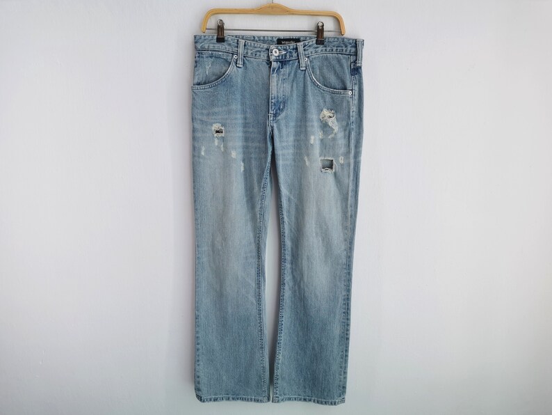 Pu&ograve; includere: Jeans in denim blu chiaro con dettagli strappati, tra cui sezioni strappate sul davanti. I jeans sono a gamba dritta e sono appesi a una gruccia di legno su sfondo bianco. Un classico capo d'abbigliamento casual.