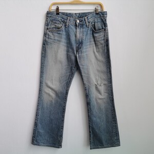 May include: Faded light blue denim jeans with a distressed knee, hanging on a wooden hanger. The jeans have a classic five-pocket design and a bootcut style. The jeans have a faded wash.