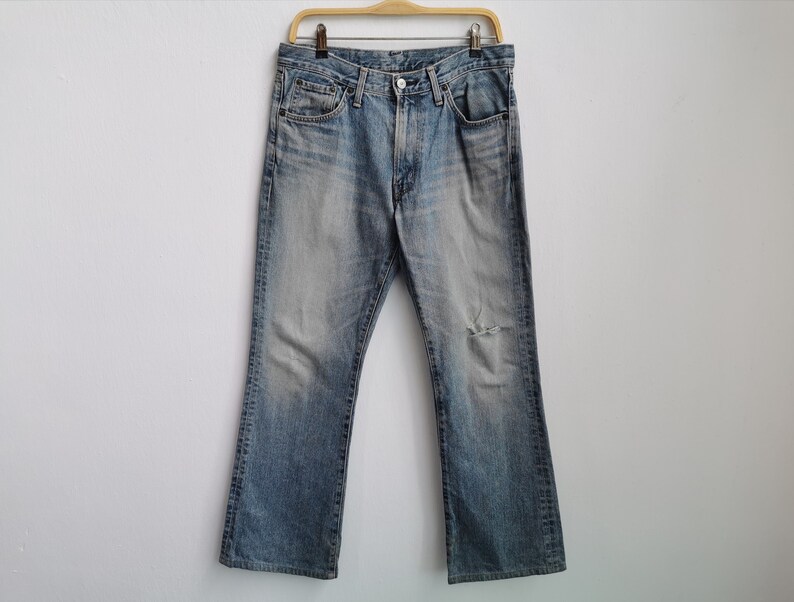 May include: Faded light blue denim jeans with a distressed knee, hanging on a wooden hanger. The jeans have a classic five-pocket design and a bootcut style. The jeans have a faded wash.