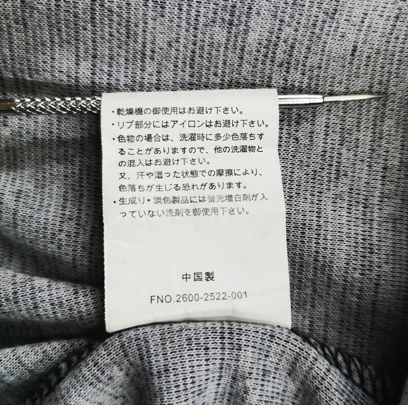 May include: Close-up of a tag with Japanese text and the text "Made in China" in black font. The tag is attached to a gray and black fabric. A silver pin is inserted through the tag.