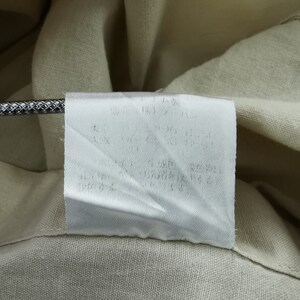 May include: A beige garment featuring a white rectangular care label sewn into the interior seam. The label contains printed text in Japanese characters. A metallic, textured cord is positioned horizontally across the top of the white fabric label.