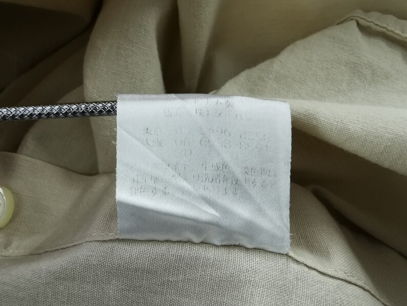 May include: A beige garment featuring a white rectangular care label sewn into the interior seam. The label contains printed text in Japanese characters. A metallic, textured cord is positioned horizontally across the top of the white fabric label.