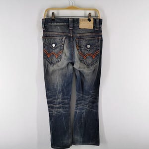 Op de afbeelding: Verbleekte blauwe denim jeans met oranje stiksels op de achterzakken. De jeans heeft een bootcut pijp en hangt aan een houten hanger. Een bruin leren label is zichtbaar op de achterkant van de tailleband.