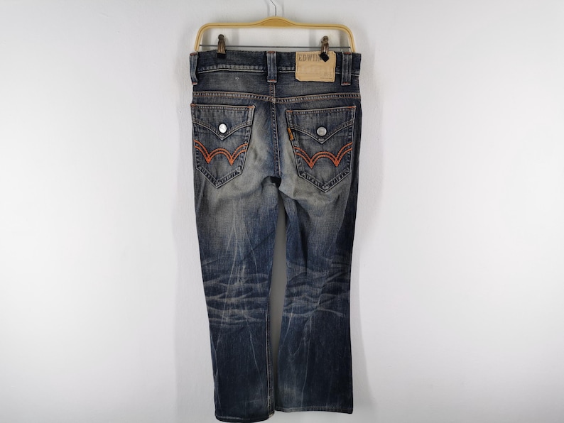 Op de afbeelding: Verbleekte blauwe denim jeans met oranje stiksels op de achterzakken. De jeans heeft een bootcut pijp en hangt aan een houten hanger. Een bruin leren label is zichtbaar op de achterkant van de tailleband.