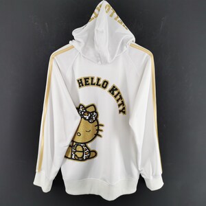 May include: White zip-up hoodie with gold stripes down the sleeves and hood. The back features a gold and black Hello Kitty graphic and the words "HELLO KITTY" in gold lettering. The hoodie is hanging against a dark gray background.