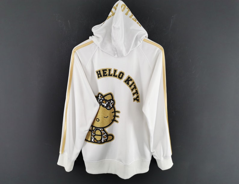 May include: White zip-up hoodie with gold stripes down the sleeves and hood. The back features a gold and black Hello Kitty graphic and the words "HELLO KITTY" in gold lettering. The hoodie is hanging against a dark gray background.