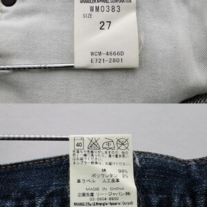 May include: Close-up of the inside of a pair of blue denim Wrangler jeans. The tags display the brand name, size 27, and care instructions. The fabric is a dark blue wash with visible stitching.