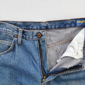 May include: Close-up of blue denim Wrangler jeans with a partially unzipped fly. The jeans have a classic five-pocket design, a button closure, and a zipper. The Wrangler label is visible on the waistband.