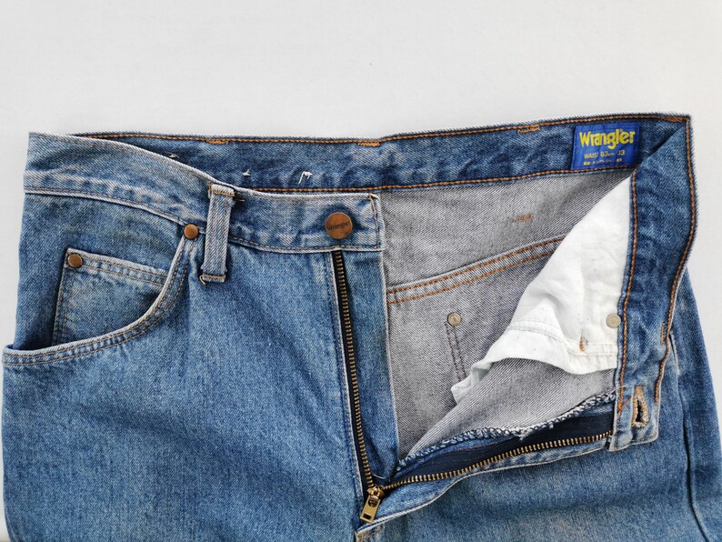 May include: Close-up of blue denim Wrangler jeans with a partially unzipped fly. The jeans have a classic five-pocket design, a button closure, and a zipper. The Wrangler label is visible on the waistband.