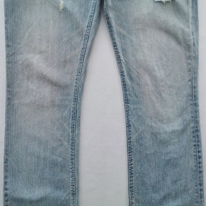 Pu&ograve; includere: Jeans in denim blu chiaro con dettagli strappati, tra cui sezioni strappate vicino alle ginocchia. I jeans hanno un classico design a cinque tasche e un taglio dritto. Il tessuto mostra sbiadimento e usura.