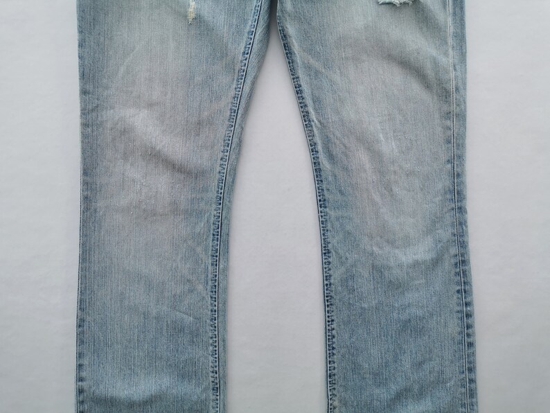 Pu&ograve; includere: Jeans in denim blu chiaro con dettagli strappati, tra cui sezioni strappate vicino alle ginocchia. I jeans hanno un classico design a cinque tasche e un taglio dritto. Il tessuto mostra sbiadimento e usura.