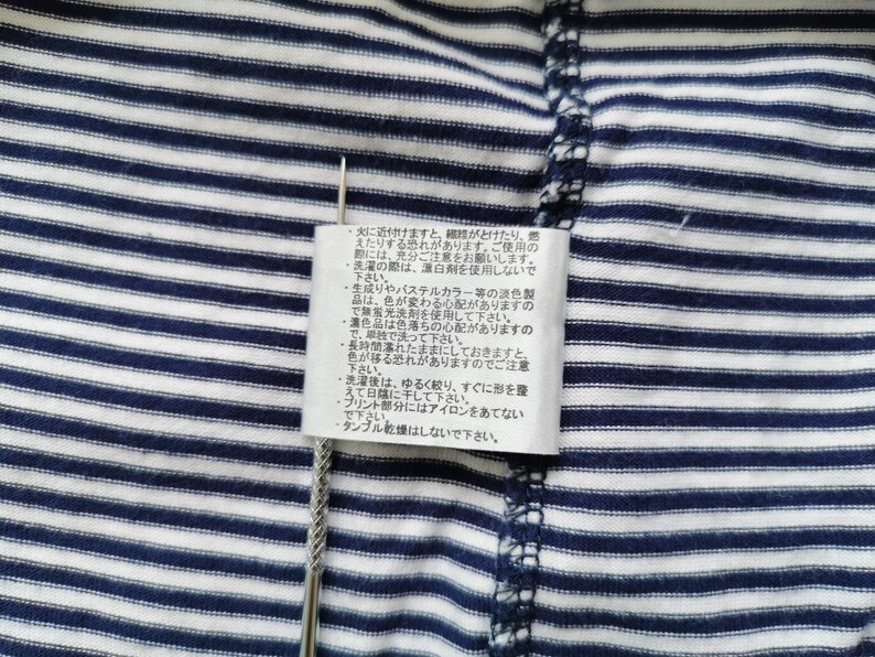 Puede incluir: Primer plano de una tela a rayas azul marino y blanca con una etiqueta de cuidado adjunta. La etiqueta contiene texto japon&eacute;s con instrucciones de cuidado. Un alfiler plateado se inserta a trav&eacute;s de la tela y la etiqueta.