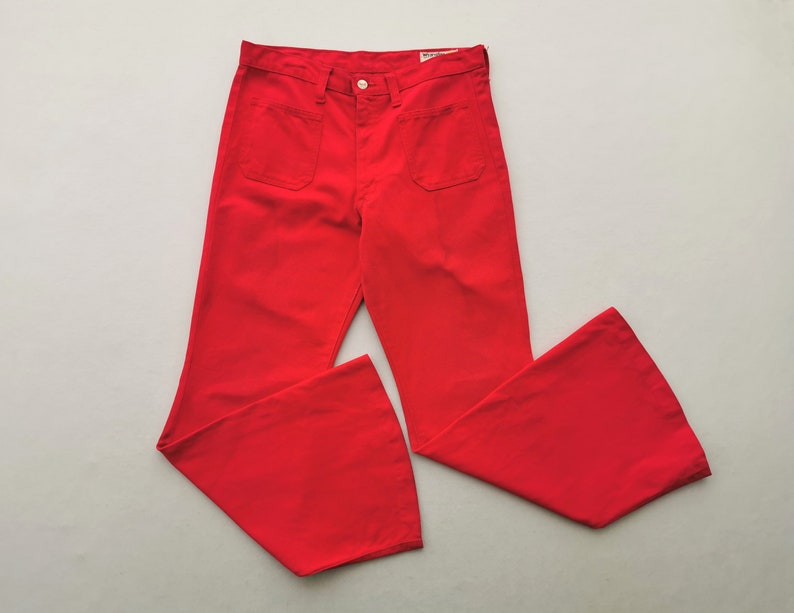 May include: Bright red flared pants with two front pockets and a button closure. The pants have a high waist and a wide leg opening. The fabric appears to be a medium-weight material. The pants are laid flat on a light gray surface.