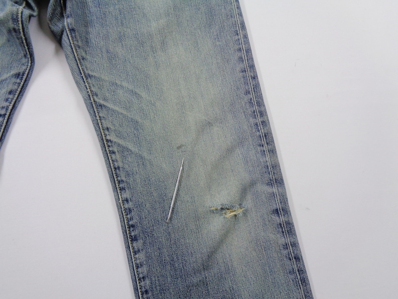 Pu&ograve; includere: Jeans di denim blu sbiadito con un aspetto usurato. I jeans presentano un aspetto vissuto con un piccolo strappo nella zona del ginocchio. Il denim ha un lavaggio chiaro e un classico design a cinque tasche.