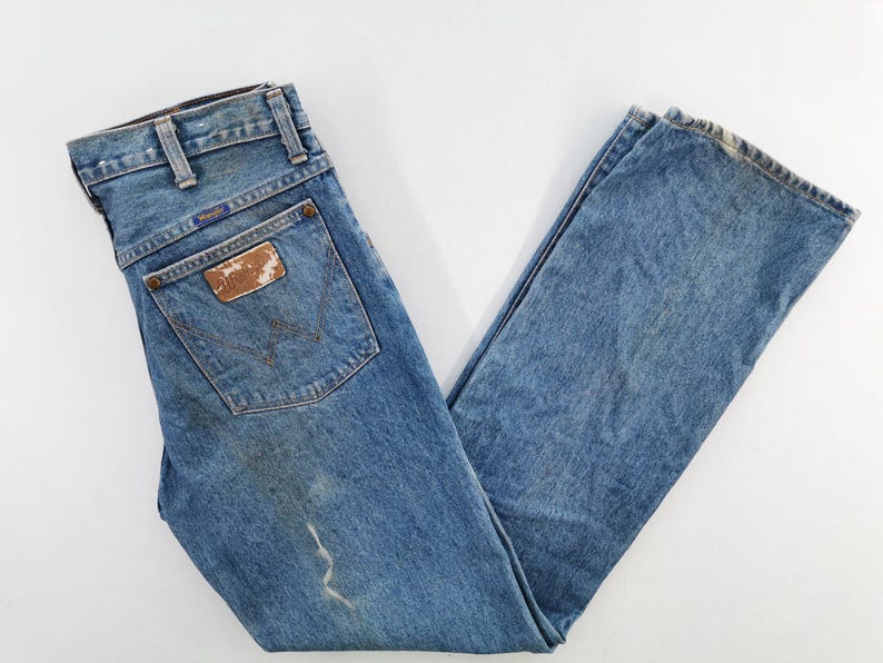 May include: Light blue denim jeans with a classic five-pocket design. The jeans feature a worn leather patch on the back pocket with the Wrangler logo. The jeans are laid flat on a white surface.