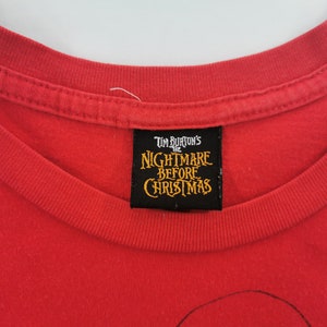 May include: Red t-shirt featuring the text "Tim Burton's The Nightmare Before Christmas" on a black label. The shirt has a crew neck and is made of a soft, likely cotton, material. The image is a close-up of the shirt's neckline and label.