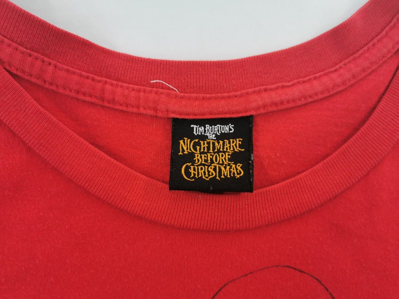 May include: Red t-shirt featuring the text "Tim Burton's The Nightmare Before Christmas" on a black label. The shirt has a crew neck and is made of a soft, likely cotton, material. The image is a close-up of the shirt's neckline and label.
