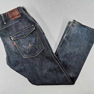 May include: Dark wash denim jeans with a brown leather patch and a red logo tag. The jeans feature a unique pocket design with decorative stitching. The denim has a faded, worn look.