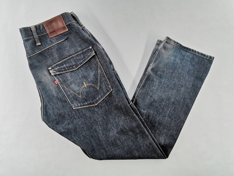 May include: Dark wash denim jeans with a brown leather patch and a red logo tag. The jeans feature a unique pocket design with decorative stitching. The denim has a faded, worn look.
