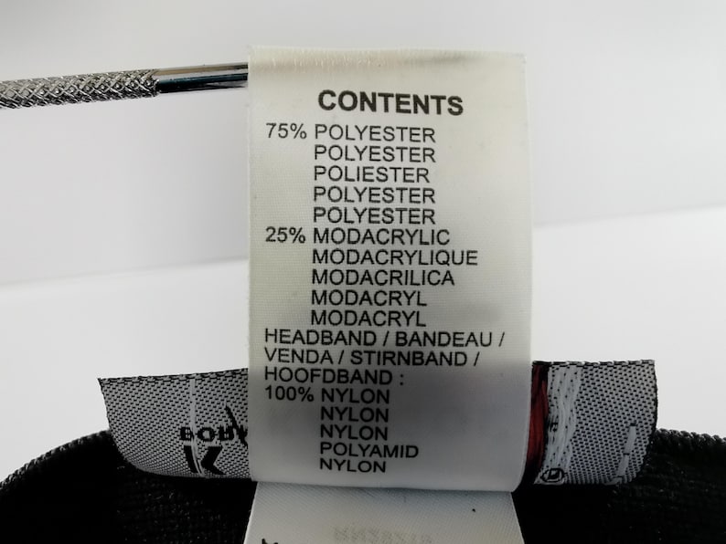 Puede incluir: Primer plano de una etiqueta blanca con texto negro que detalla la composici&oacute;n de una diadema. La etiqueta enumera materiales como poli&eacute;ster, modacr&iacute;lico y nailon. La palabra "HEADBAND" tambi&eacute;n est&aacute; escrita en varios idiomas.